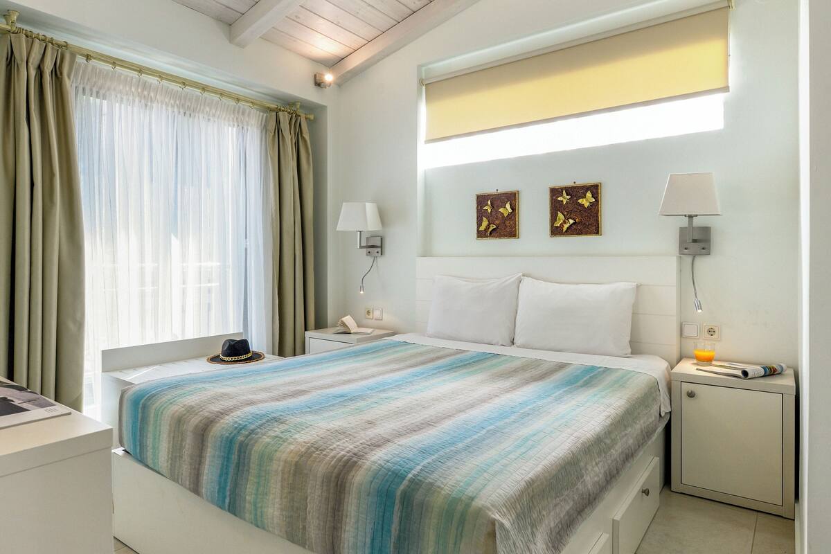 Une chambre à coucher comprenant un lit, deux tables de chevet, une commode, une fenêtre avec des rideaux et un plafond avec des poutres en bois.