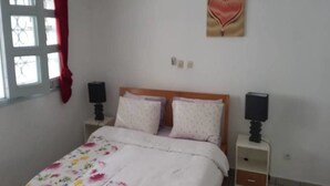 A bedroom with a floral bedspread, a heart-shaped wall decoration, two bedside tables with lamps, and a window with a red curtain. - Résidence Petite Villa (Abidjan)