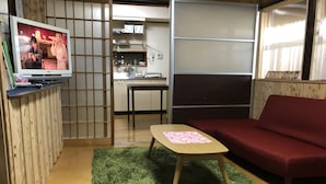 Ett kompakt vardagsrum med en tv som visar ett program, en röd soffa, ett litet träbord med en rosa bordsduk och en grön matta.
