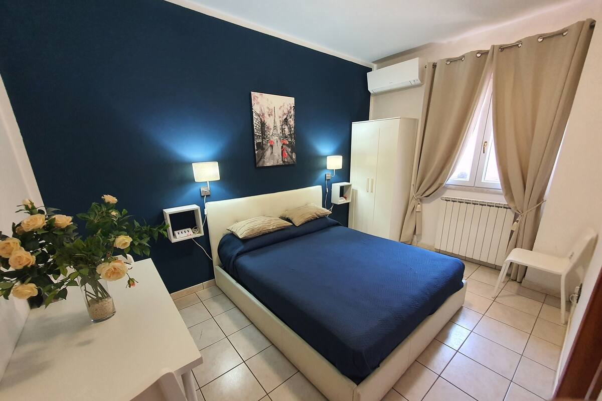 Une chambre à coucher comprenant un lit, des tables de chevet, un bureau, une chaise, un radiateur et une fenêtre avec des rideaux.