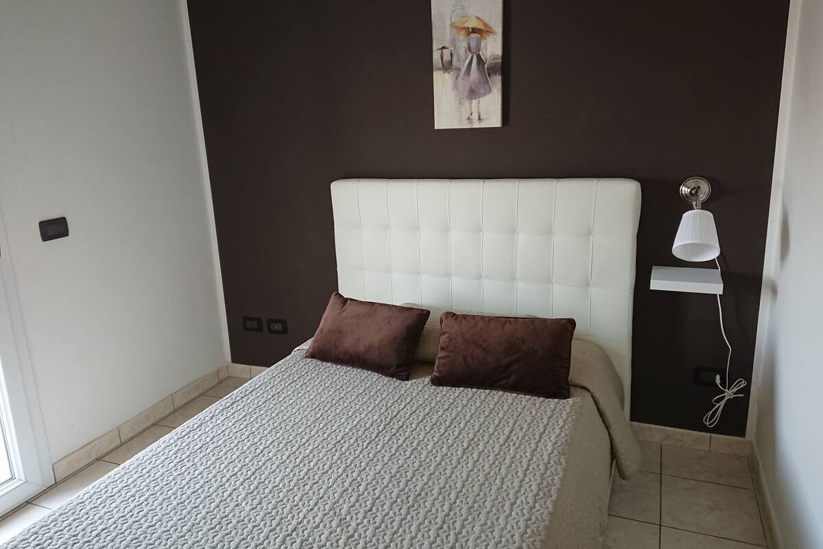 Une chambre à coucher avec un lit blanc, des oreillers marron, une tête de lit blanche, une lampe fixée au mur et un tableau.