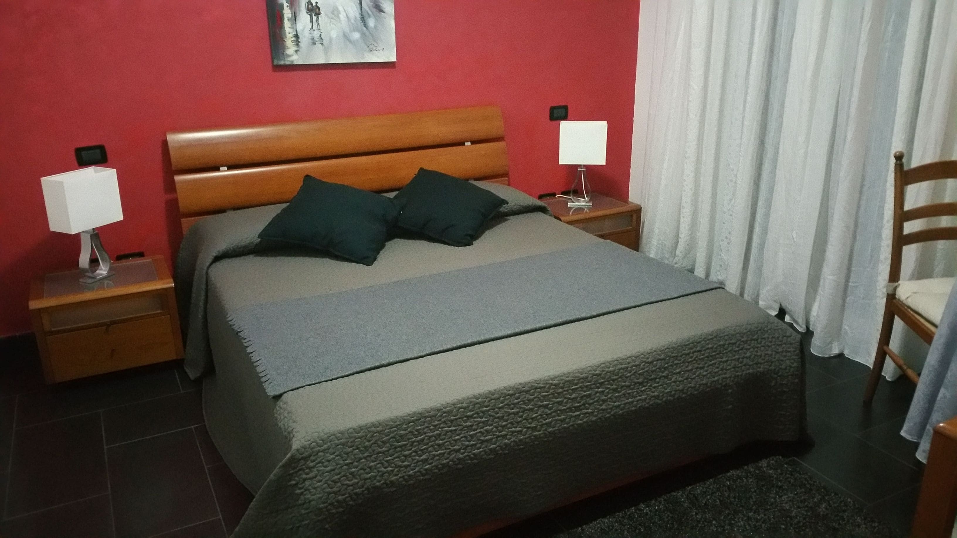 A bedroom with a wooden headboard, two bedside tables with lamps, a bed with a grey blanket and two pillows, a small nightstand, and a chair.