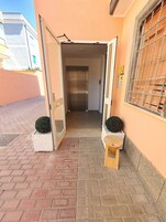 Ingresso con porta che conduce a un ascensore, un piccolo sgabello di legno e due piante in vaso.