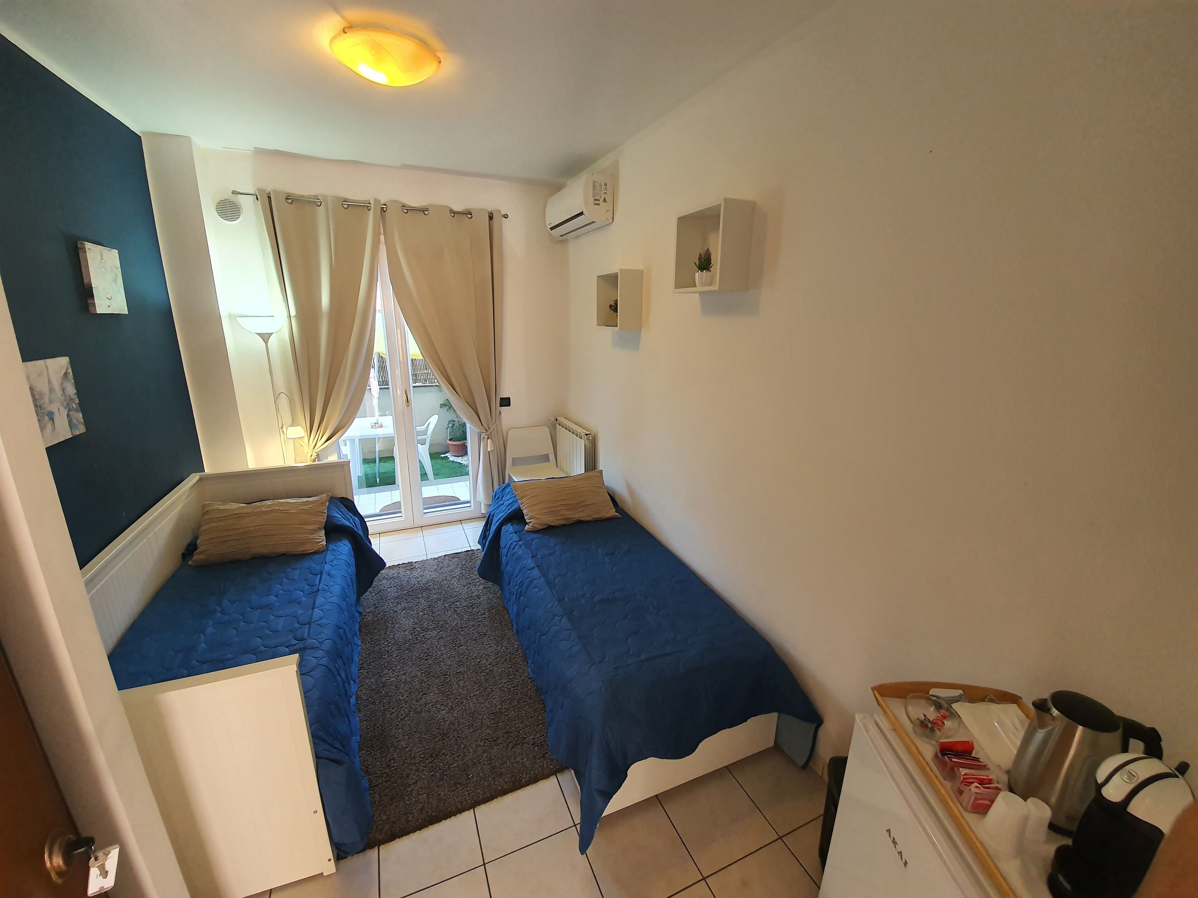 A small, well-lit room with two beds, a small table, and a kitchenette.