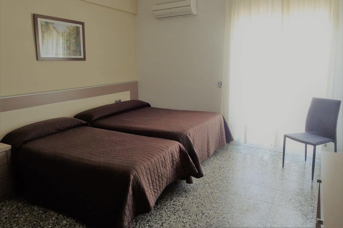 A hotel room with two single beds, a chair, an air conditioning unit, and a painting on the wall.