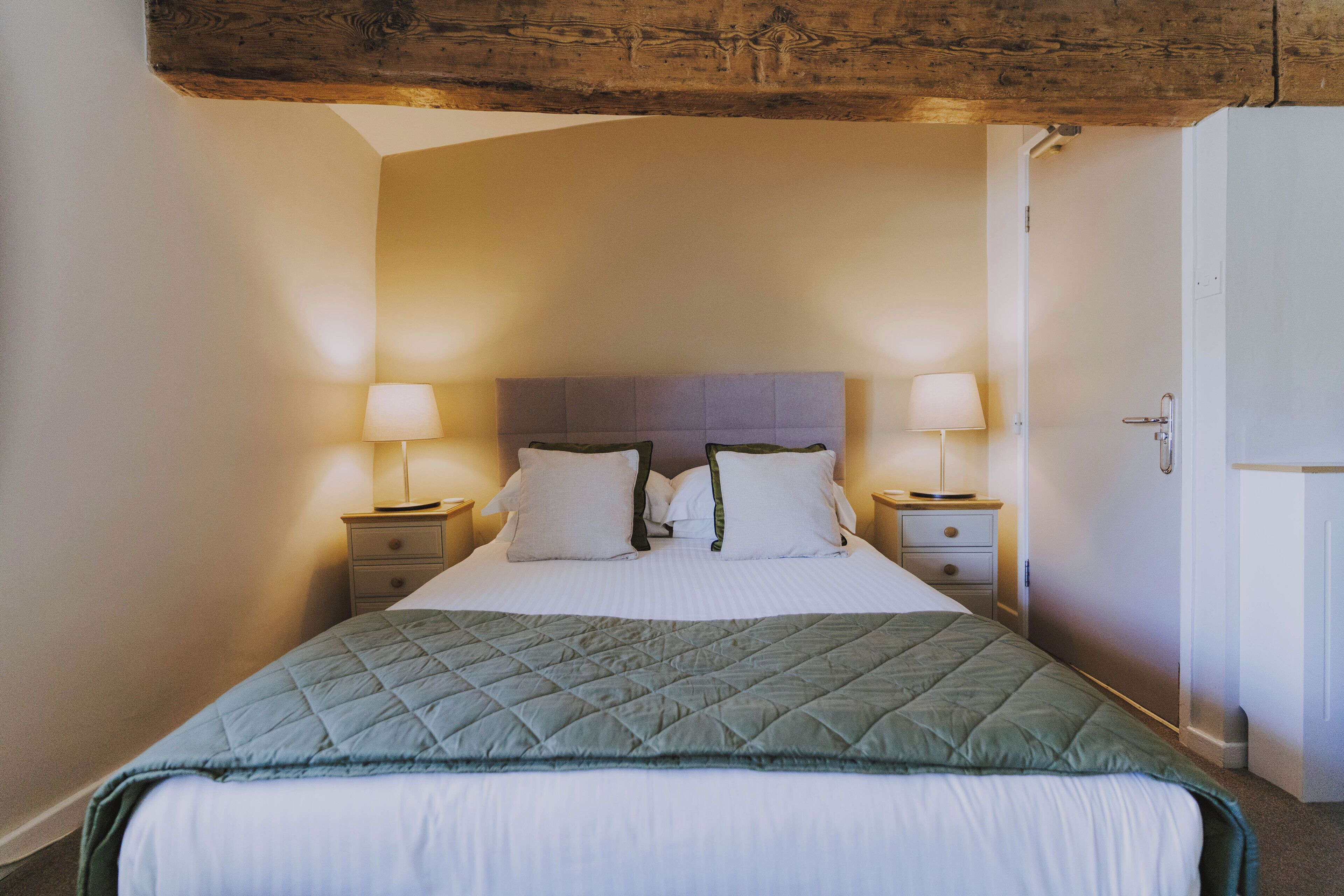 A neatly made bed with a quilted green blanket, two bedside lamps, and a wooden ceiling beam.