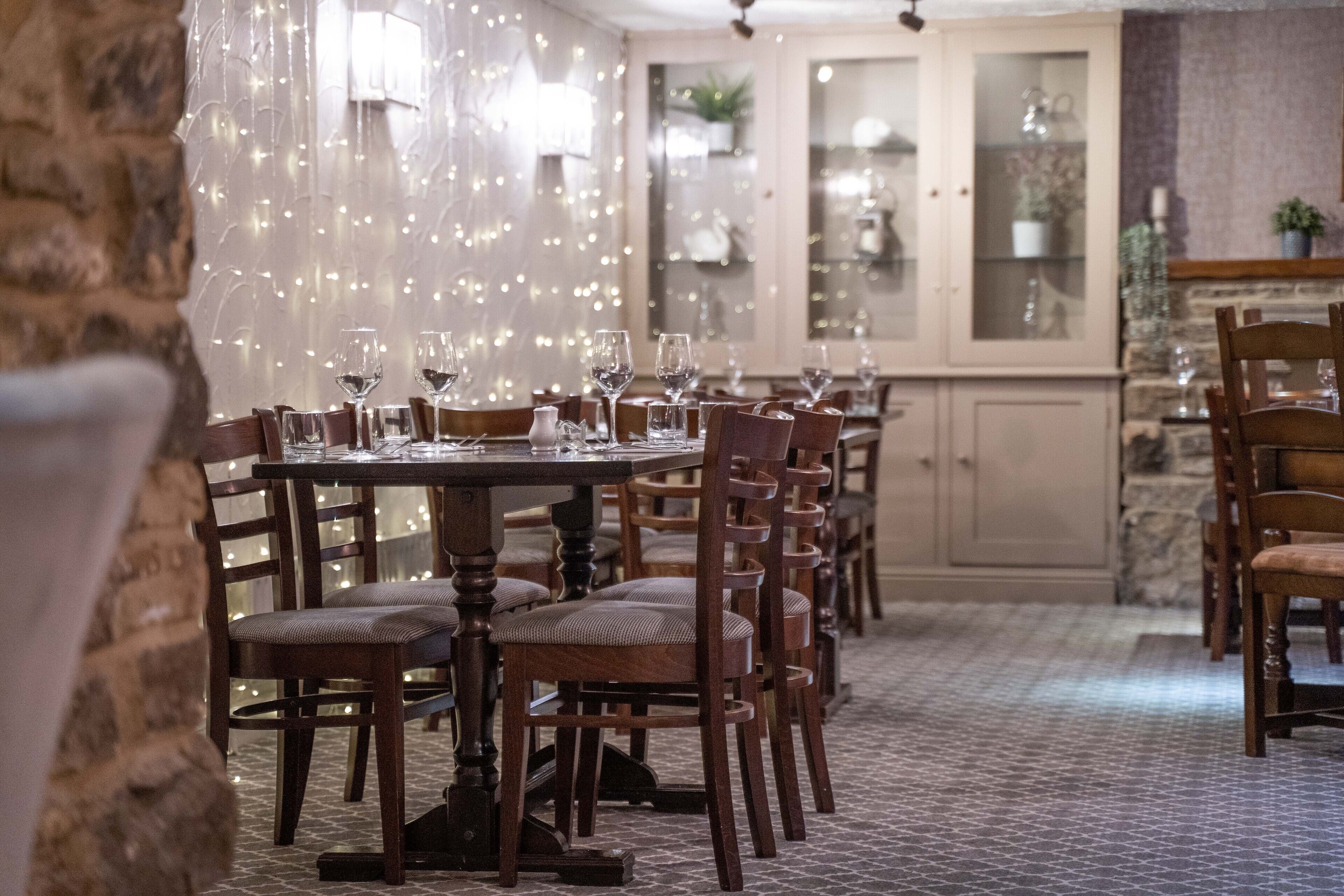 A dining area with wooden chairs and tables, string lights, and a stone wall.