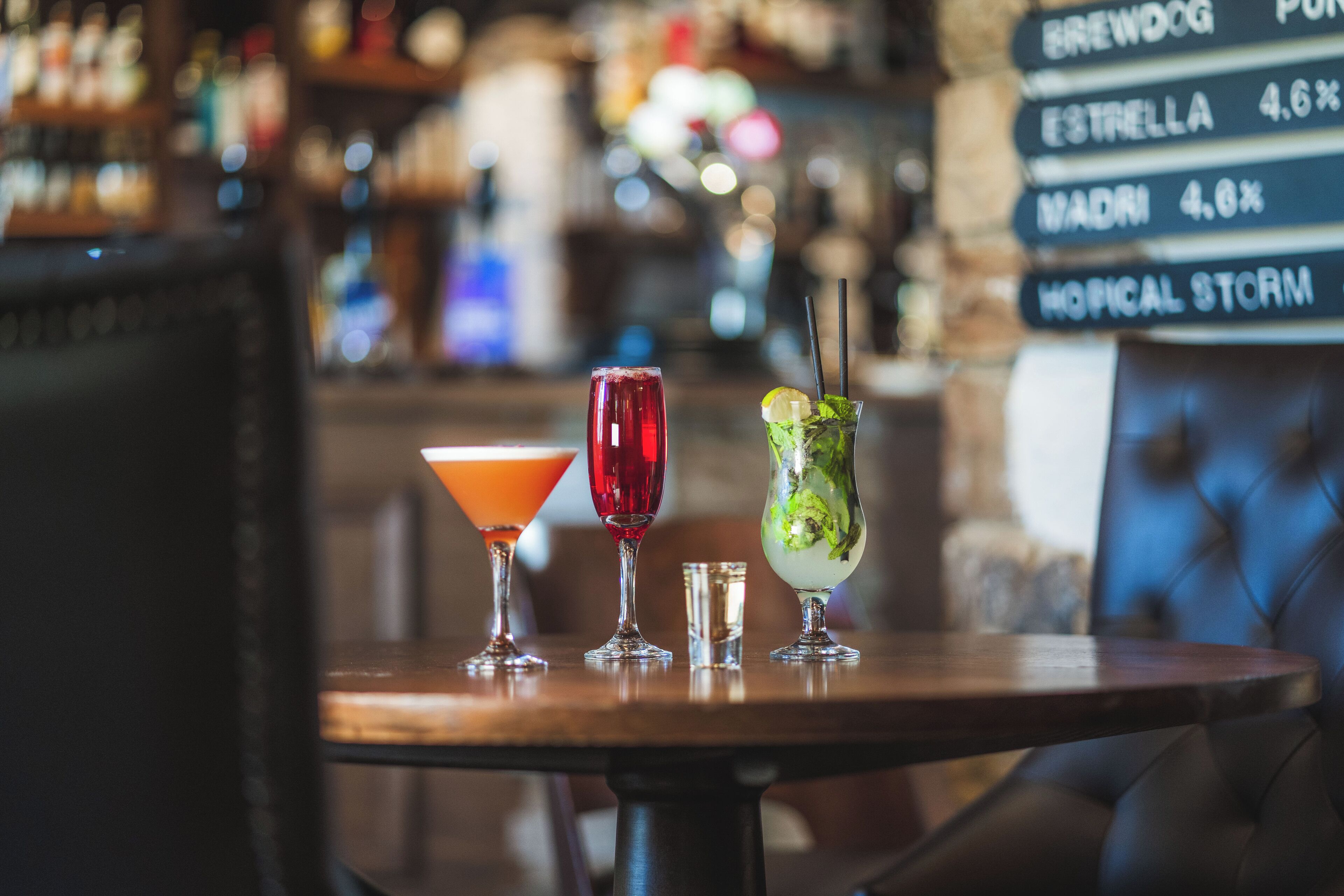 Una zona de bar con una mesa de madera que muestra tres bebidas, un panel con la carta de menú al fondo y una silla tapizada en azul.