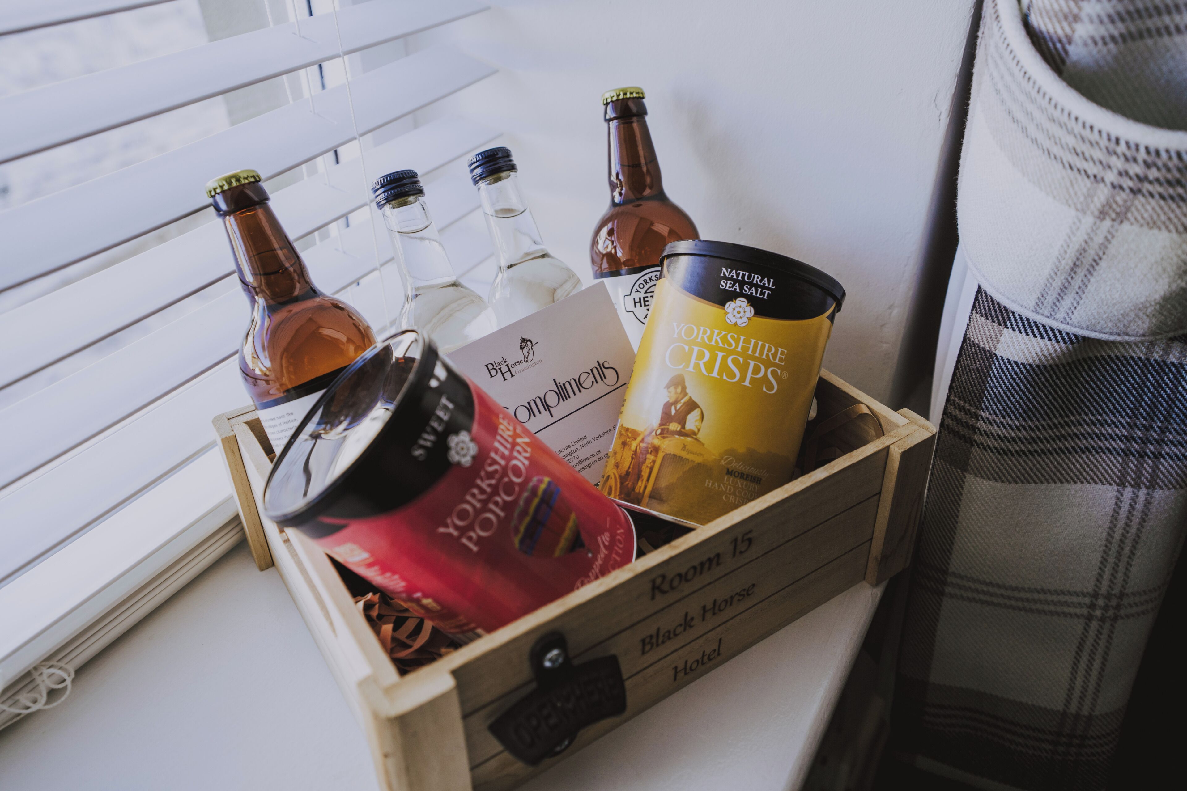 A wooden crate containing a Yorkshire crisps tin, a bottle of Black Horse beer, and a bottle of Yorkshire Peroni beer, placed on a windowsill.