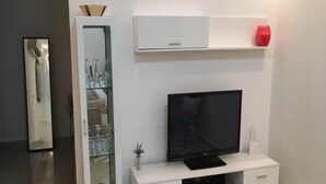 A modern living room with a flat-screen TV on a white entertainment unit, a glass cabinet with decorative items, and a red vase on a shelf. - Résidence Lumière 3 (Abidjan)