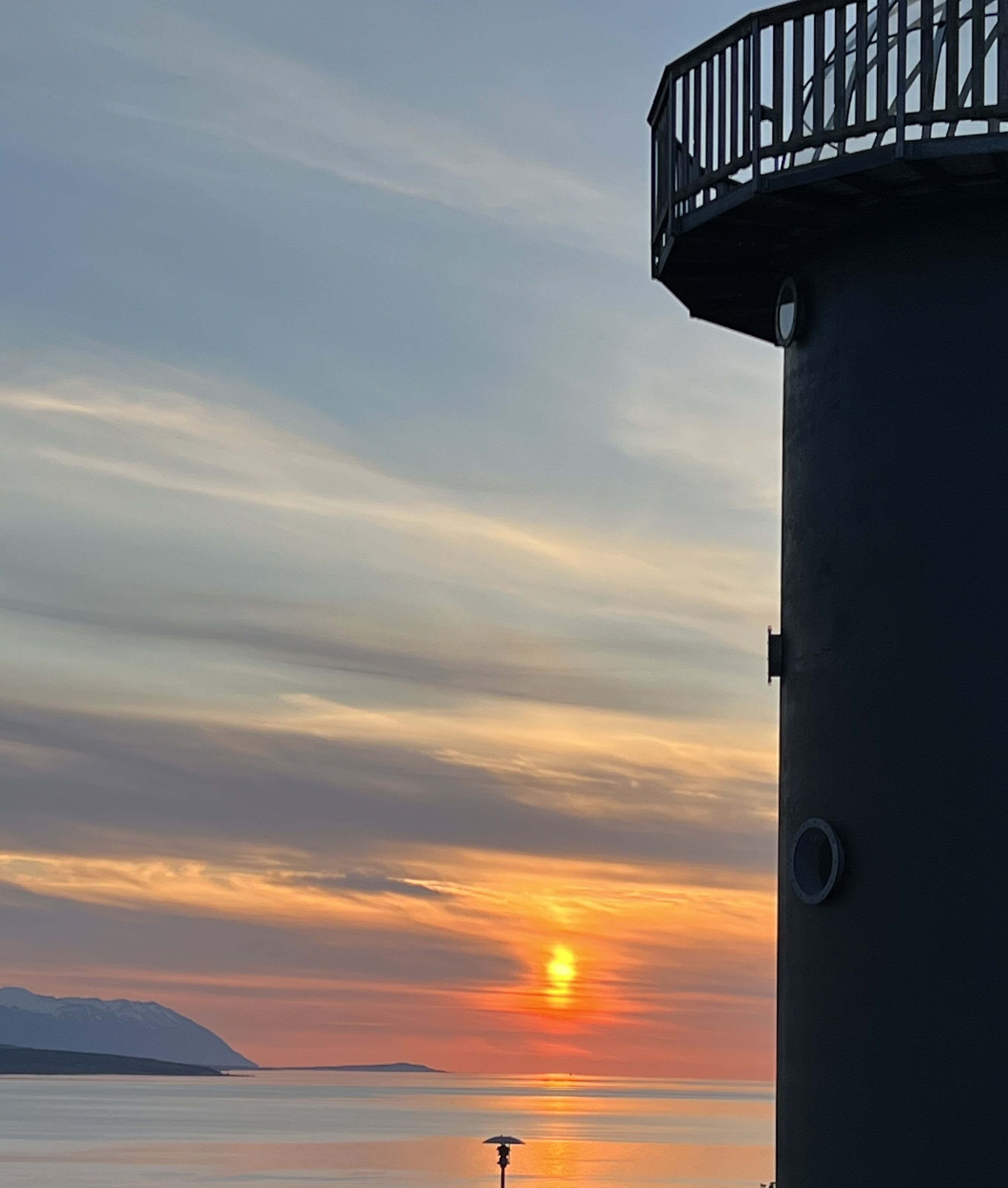 A lighthouse with a circular top and a balcony, overlooking a calm body of water during sunset.