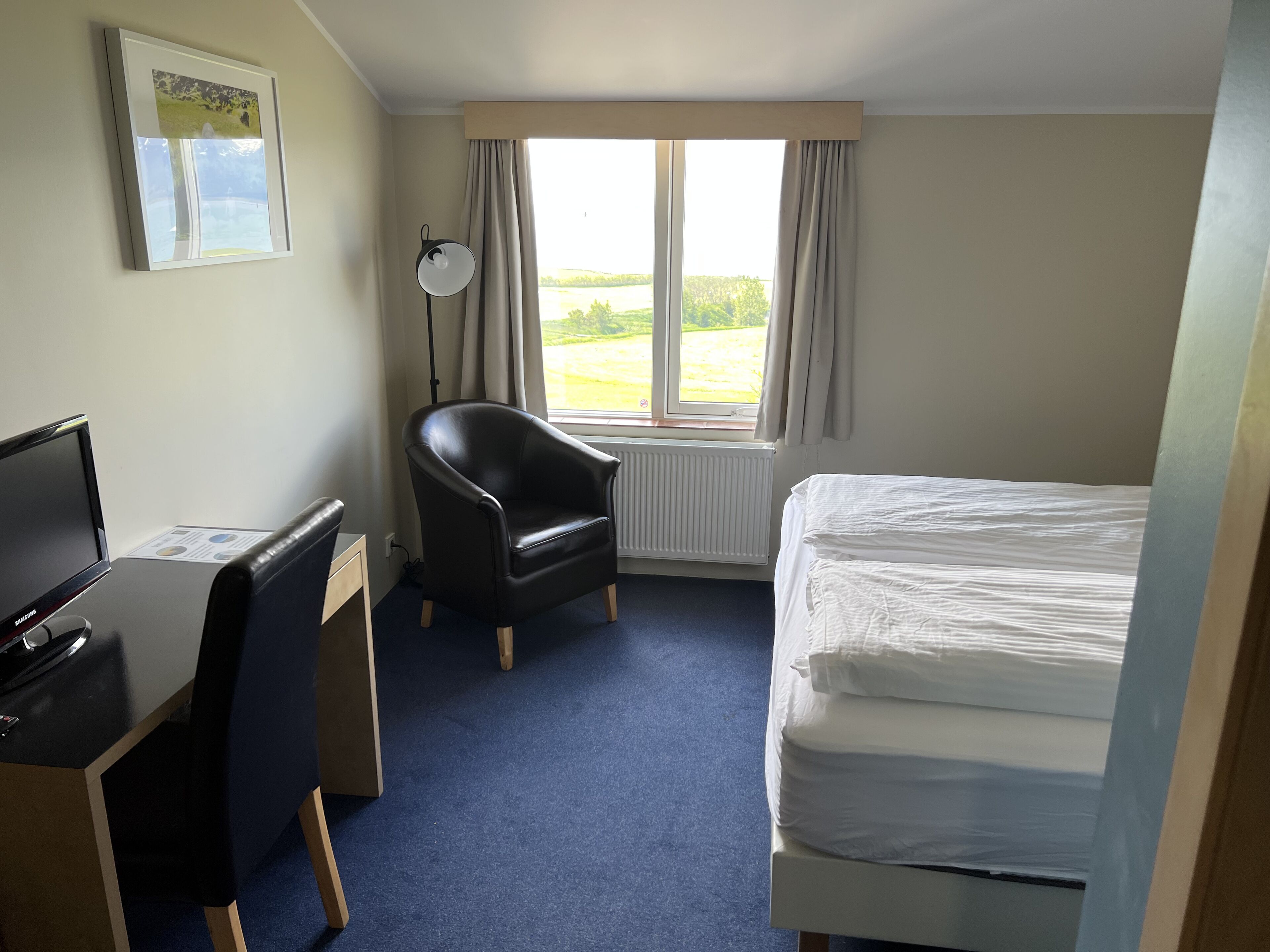 A hotel room with a bed, a desk with a computer, a chair, and a window with curtains.