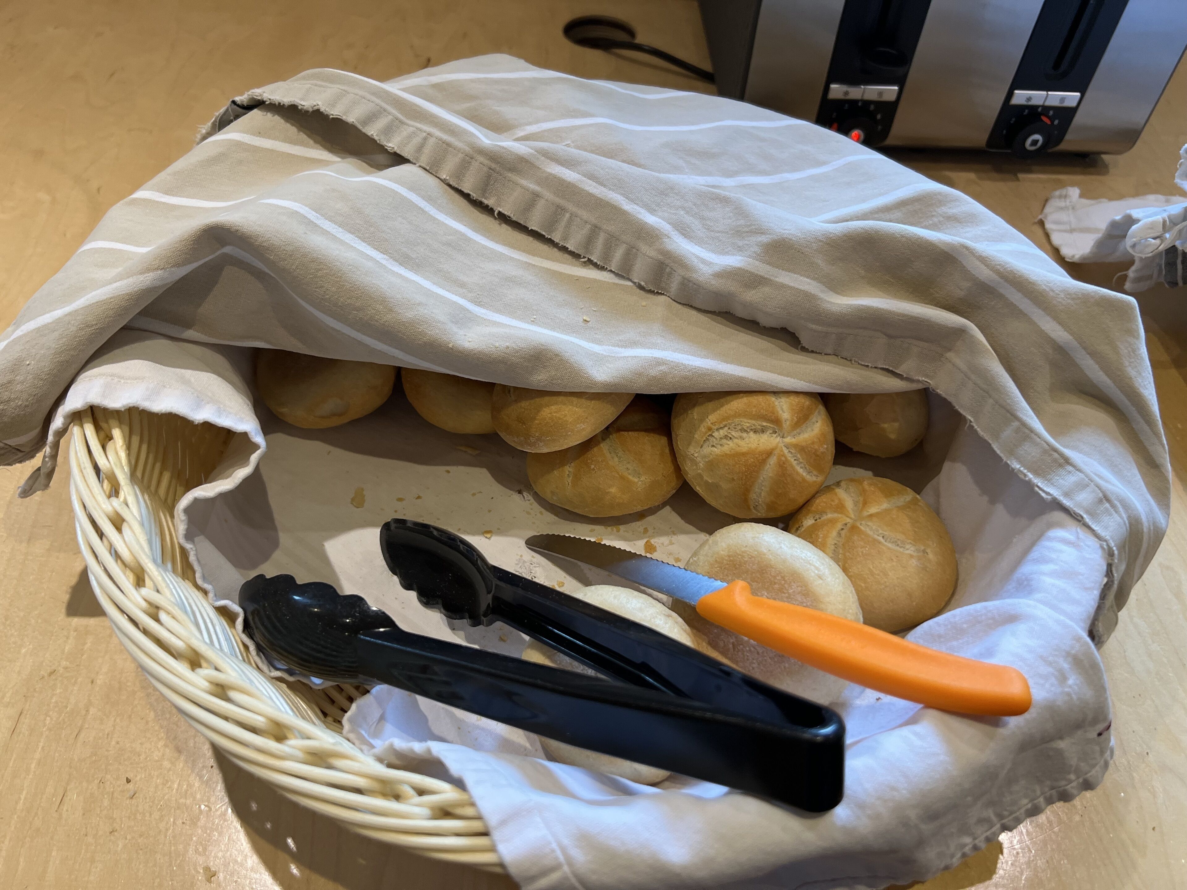 A basket of bread rolls on a wooden table with a bread knife and tongs.