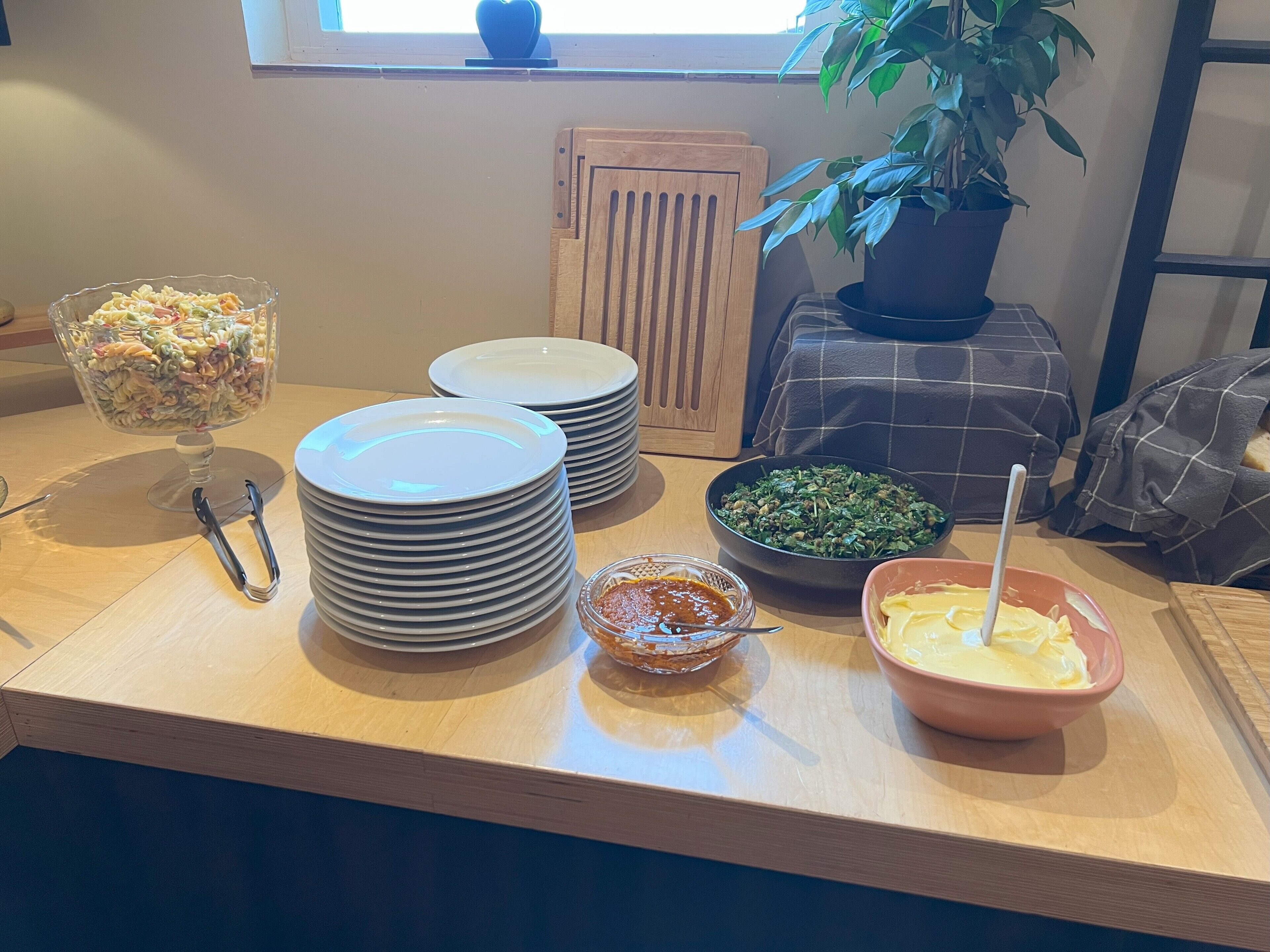 Una mesa de madera con un bol de ensalada de pasta, pilas de platos blancos, un bol de vegetales verdes, un bol de salsa y un bol de aderezo.