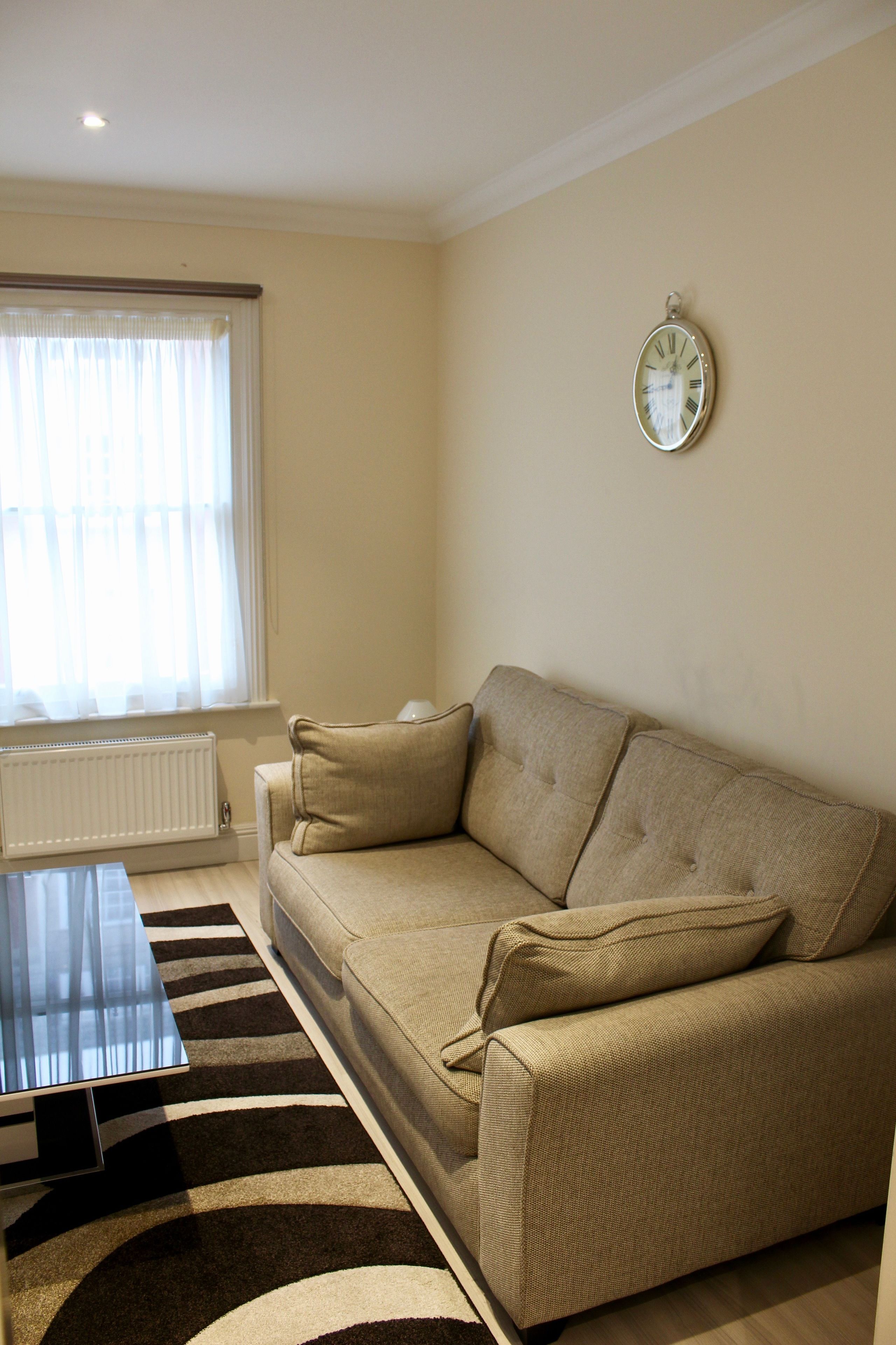 A living room with a beige sofa, a coffee table, a rug, a window with curtains, and a wall clock.