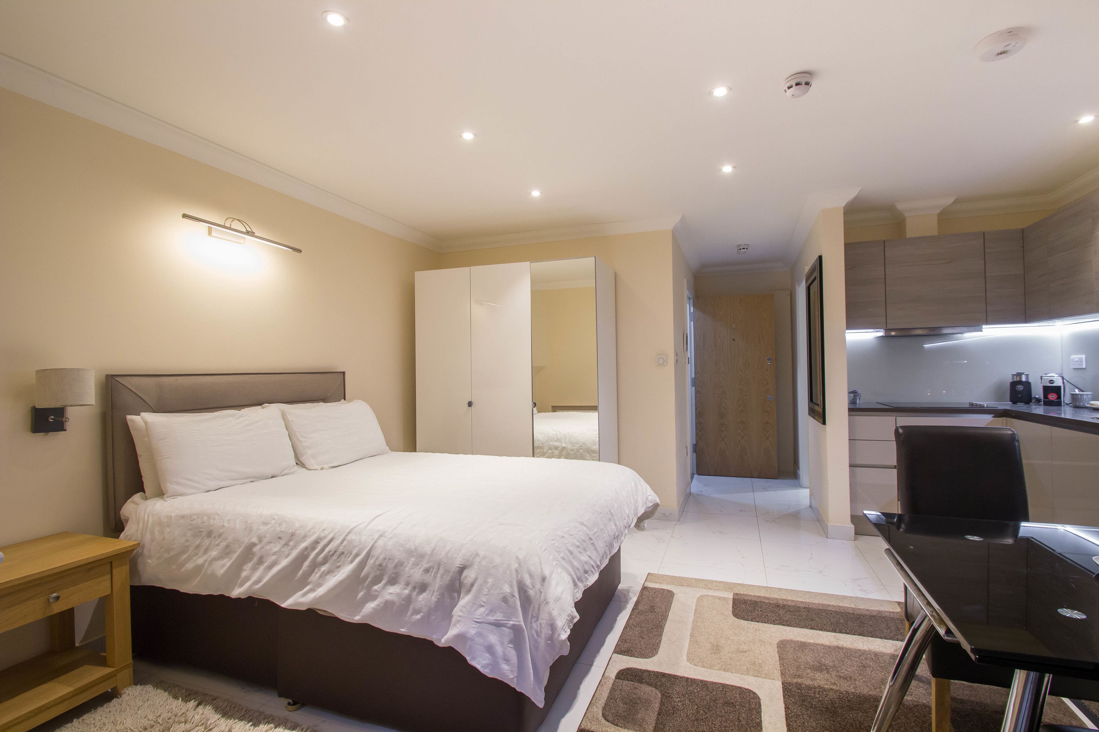 A bedroom with a large bed, bedside table, and a desk with a chair. There is a kitchenette area with cabinets and a countertop.