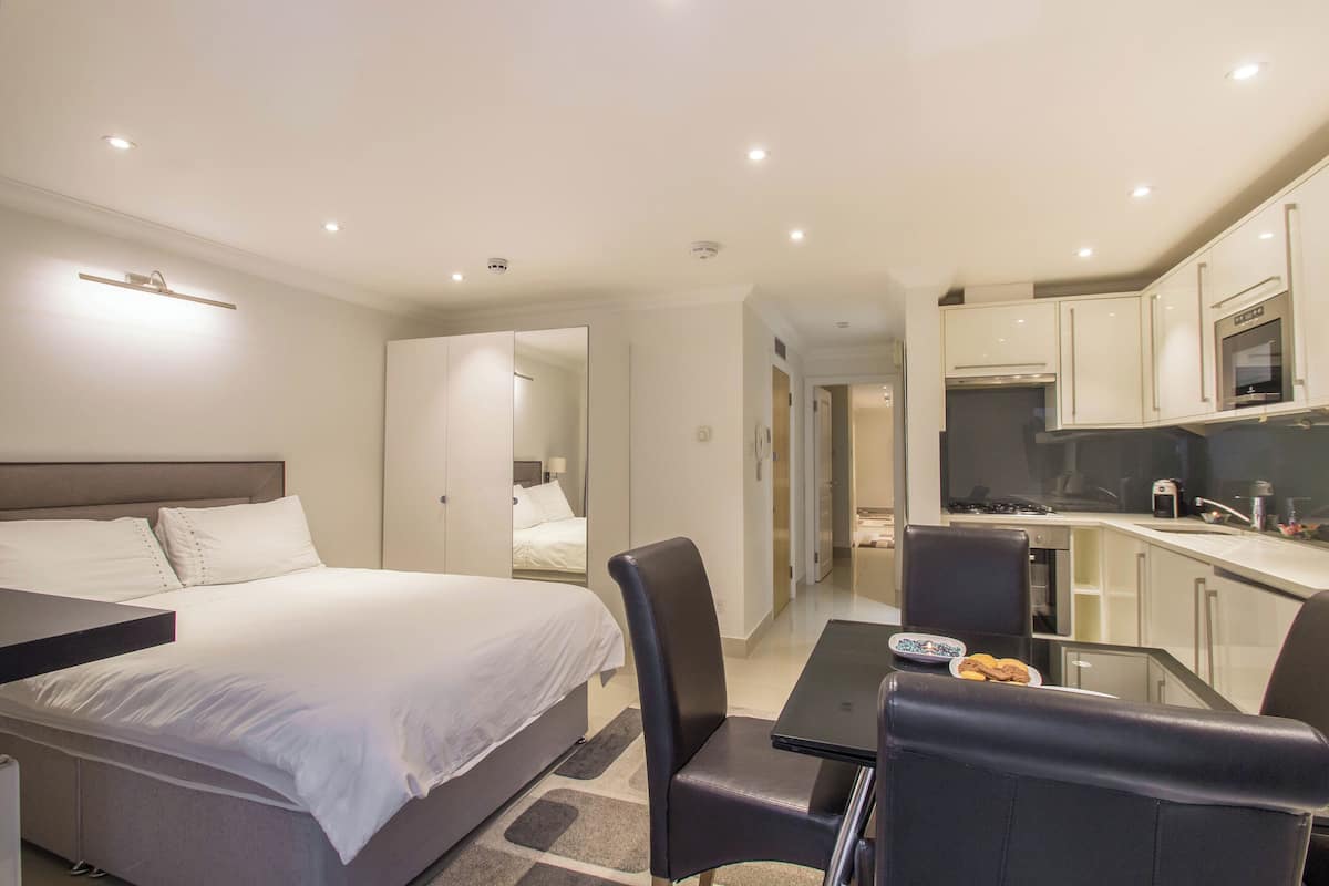 A modern hotel room with a bed, a desk, a chair, a small table with a plate of cookies, and a kitchenette with cabinets and a microwave.