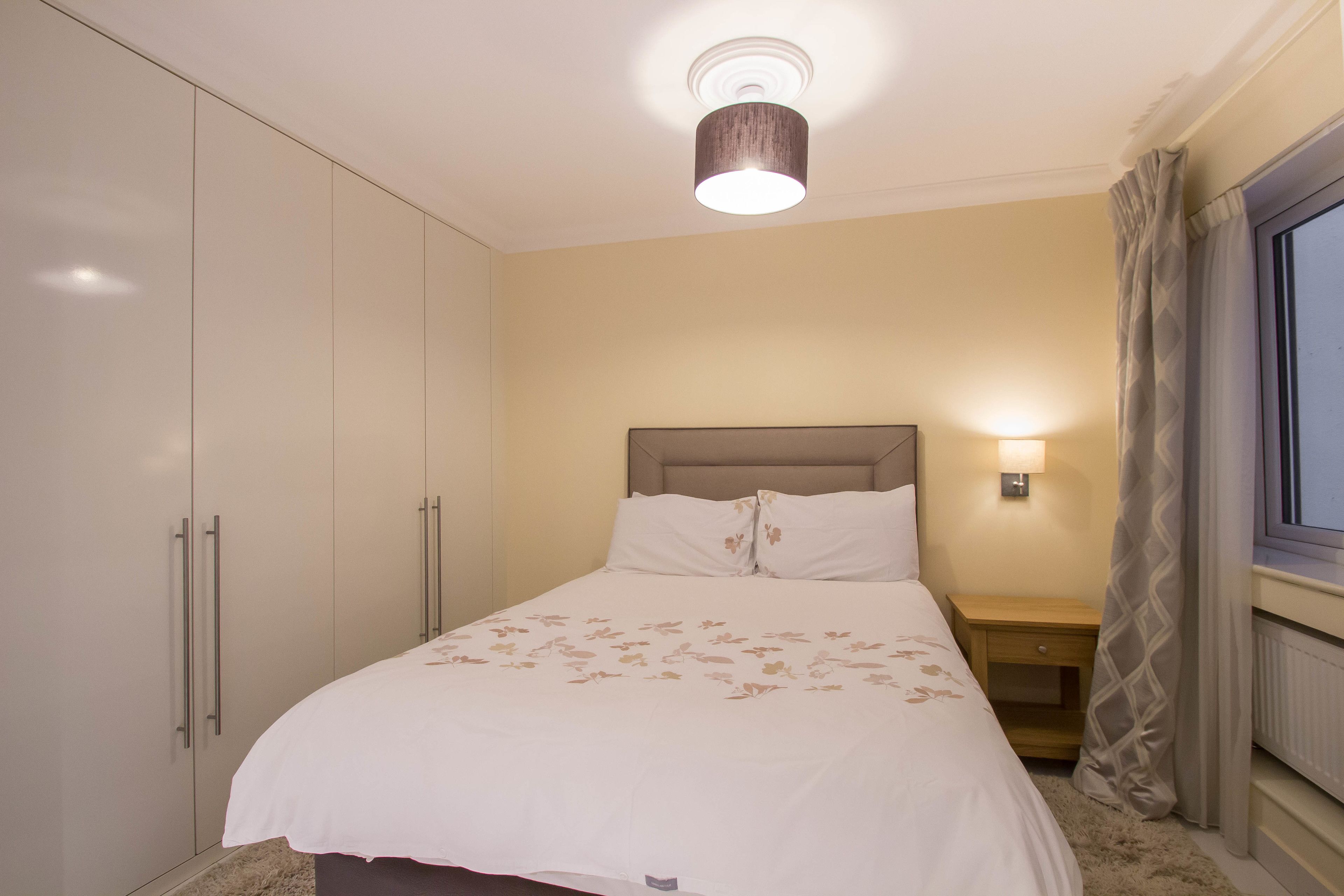 A bedroom with a bed, a nightstand, a lamp, a wardrobe, and a window with curtains.