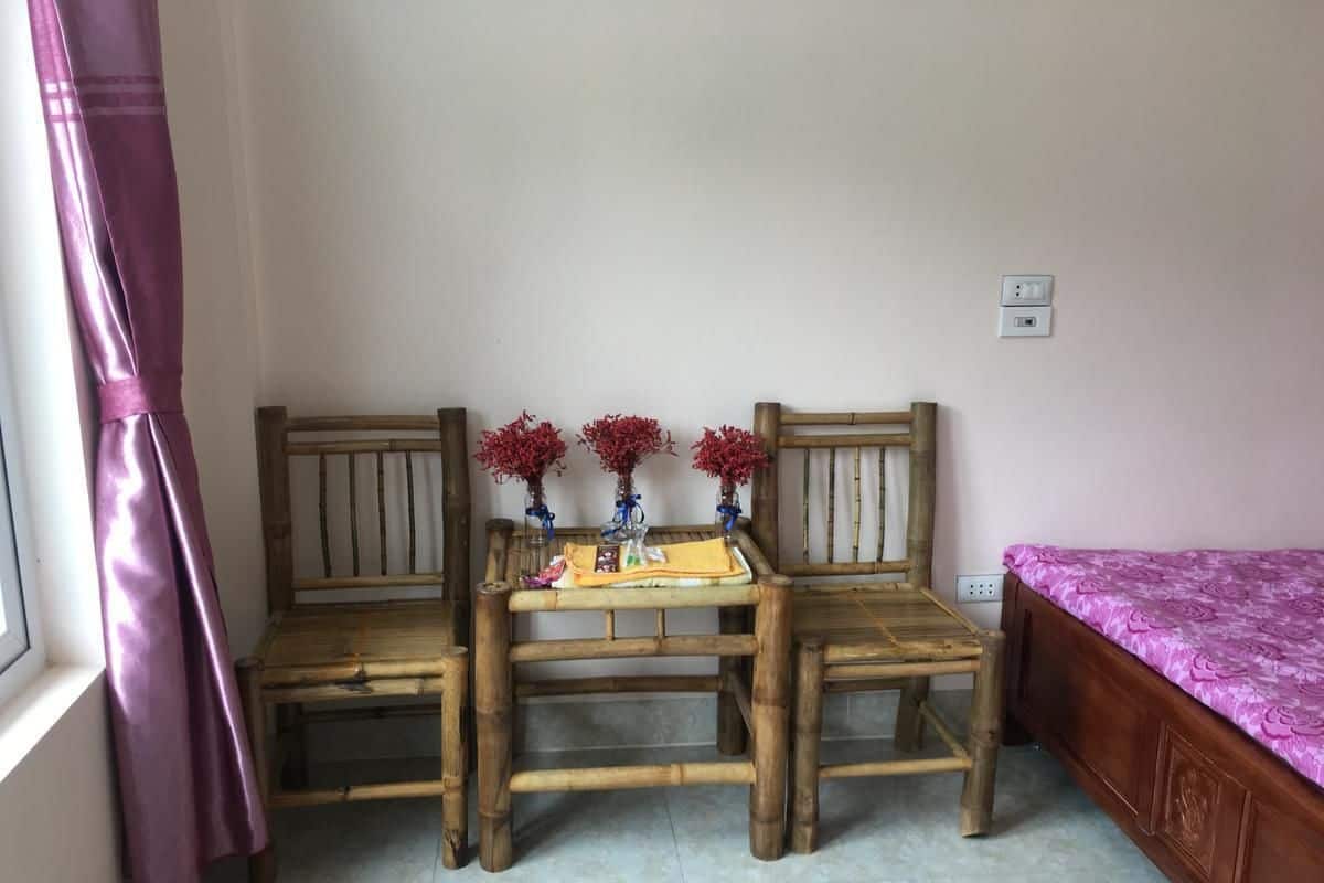 A room with two wooden chairs, a small table with a yellow cloth, and a bed with a purple bedspread.