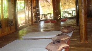 A room with wooden floors, a bed with a white mattress and two pillows, a ladder, and a view of the outdoors through large windows.
