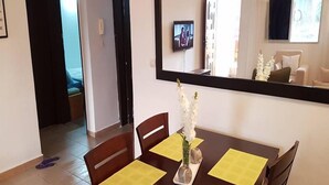A dining area with a wooden table, yellow placemats, and a vase with white flowers. A television is mounted on the wall, and a bed is visible in the background. - Résidence Marié Majeur (Abidjan)