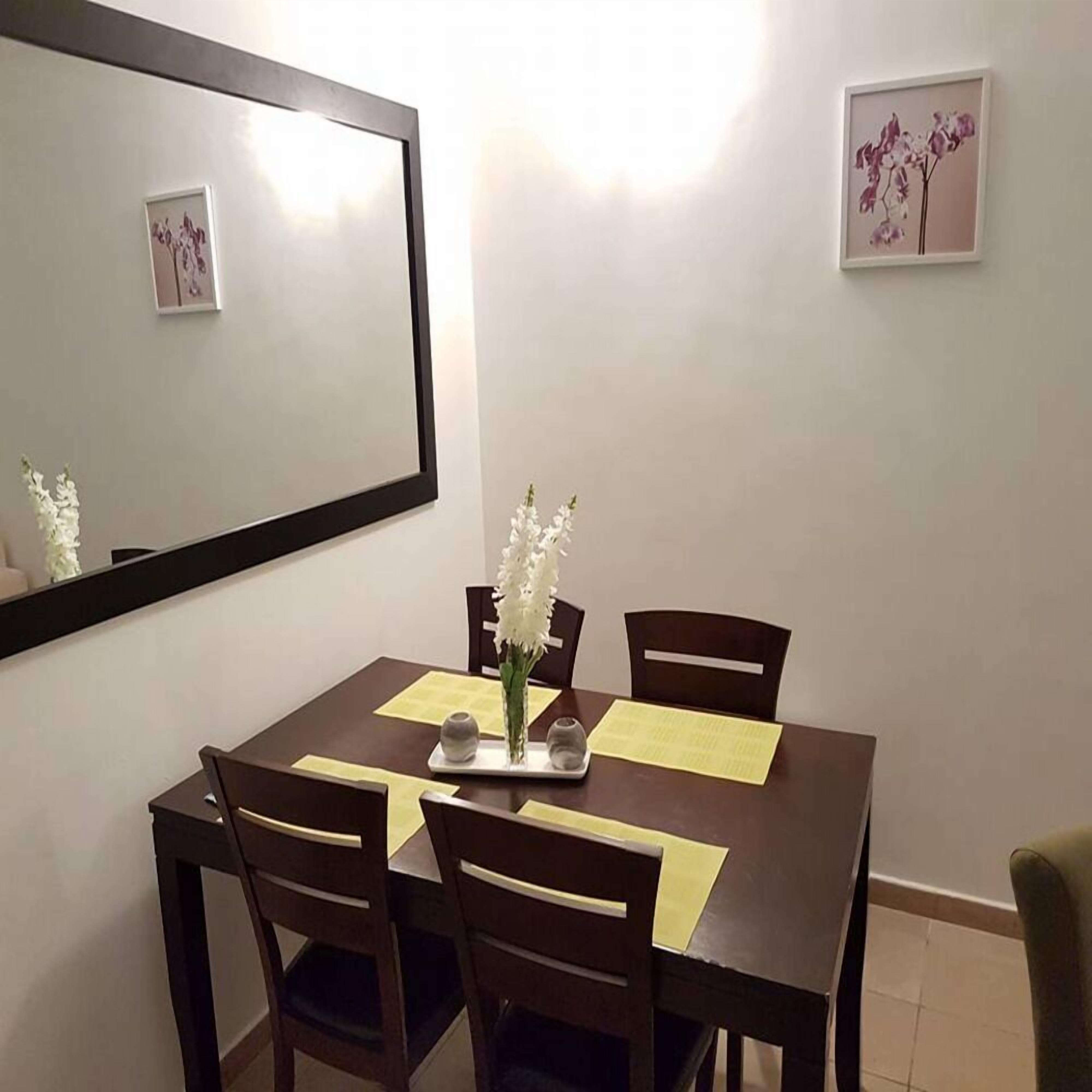 A dining area with a rectangular table, four chairs, a vase with white flowers, and a framed picture on the wall.