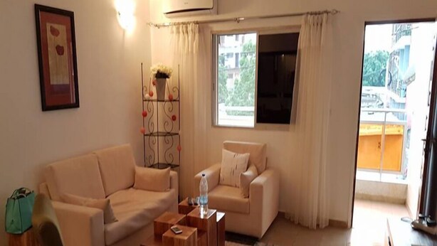 A living room with a beige sofa, a smaller armchair, a wooden coffee table, and a large window with curtains. - Résidence Marié Majeur (Abidjan)