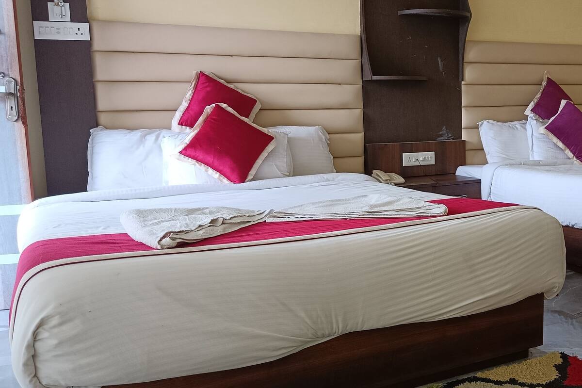 A hotel room with two beds, each with white bedding and red and purple pillows. There is a wooden headboard, a bedside table with a phone, and a wall socket.