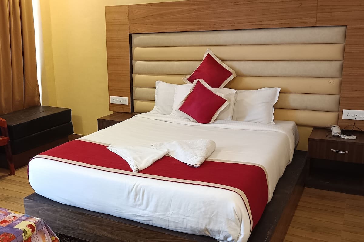 A hotel room with a large bed, a wooden headboard, a nightstand, a chair, and a table with a colorful tablecloth.