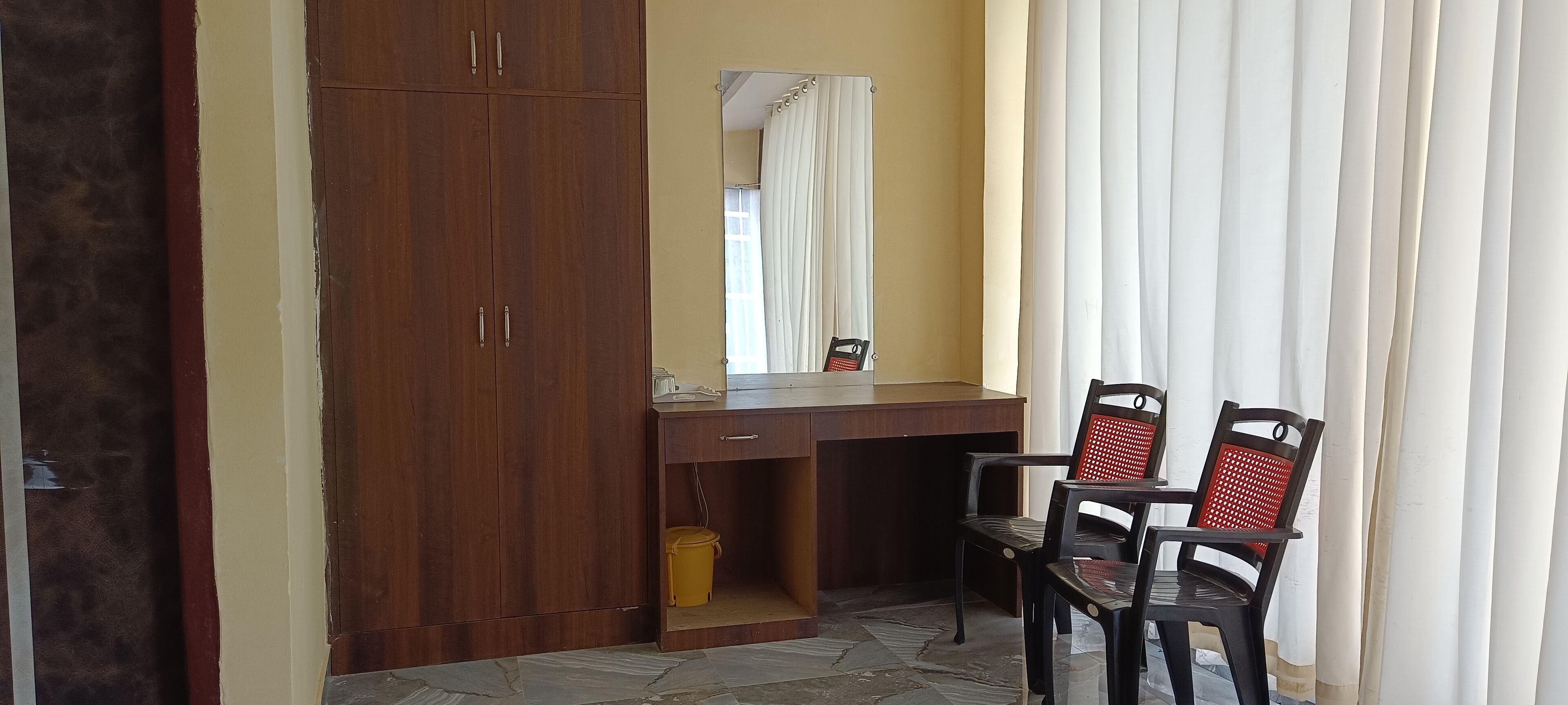 Ein Zimmer mit einem Holzschrank, einem Schreibtisch mit Spiegel, zwei Stühlen und einem gelben Mülleimer.