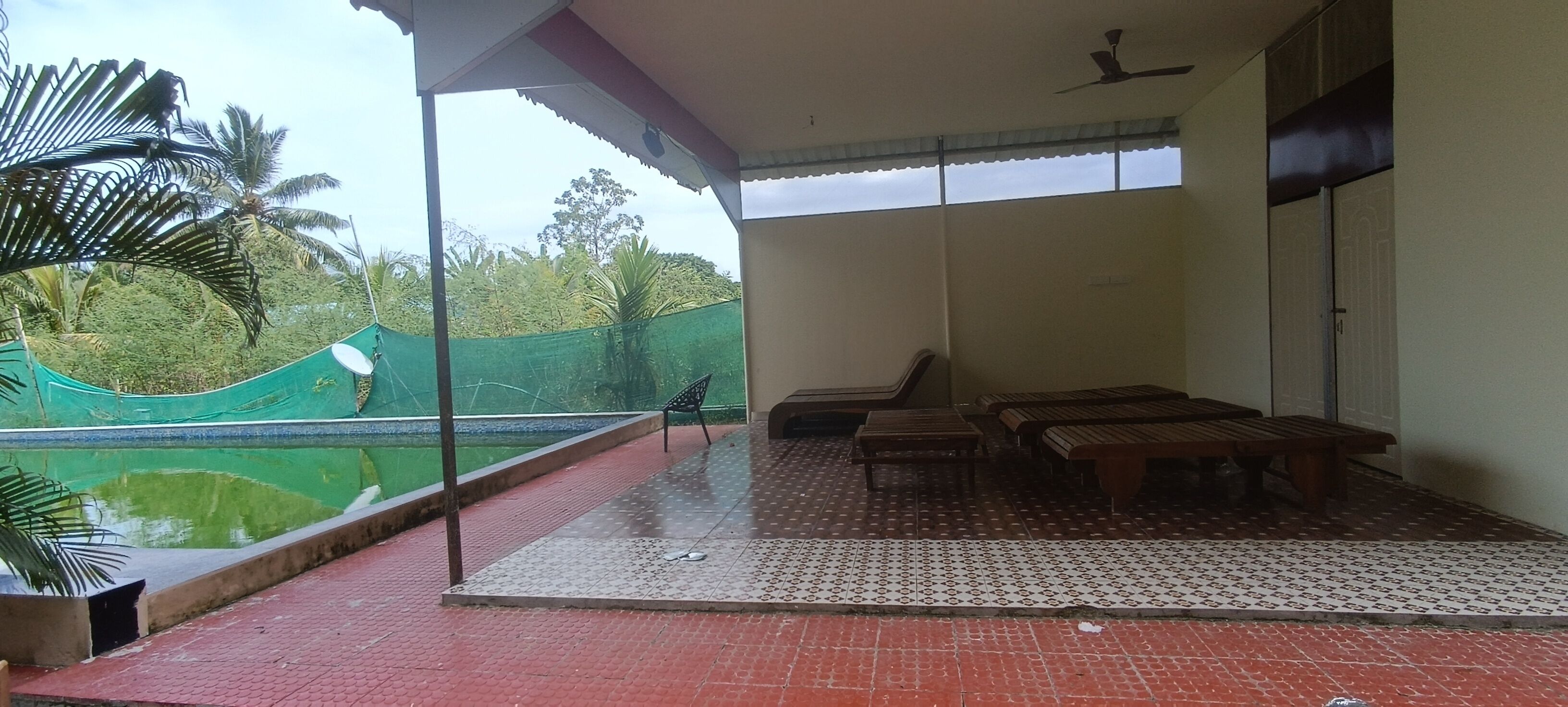 Un balcon abrité offrant une vue sur une piscine et des palmiers, avec des bancs en bois et un ventilateur de plafond.
