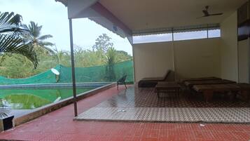 Un balcón techado con vista a una piscina y palmeras, bancos de madera y un ventilador de techo.