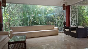 Um lobby moderno com um sofá bege, uma mesa de centro de vidro, mobiliário de vime e vista para vegetaç ão exuberante através de um painel decorativo de metal.