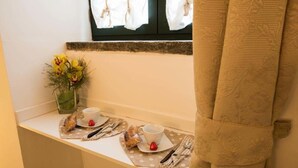 Un asiento junto a una pequeña ventana con dos tazas y platillos blancos, un florero de cristal con flores amarillas y una bolsa de aperitivos.