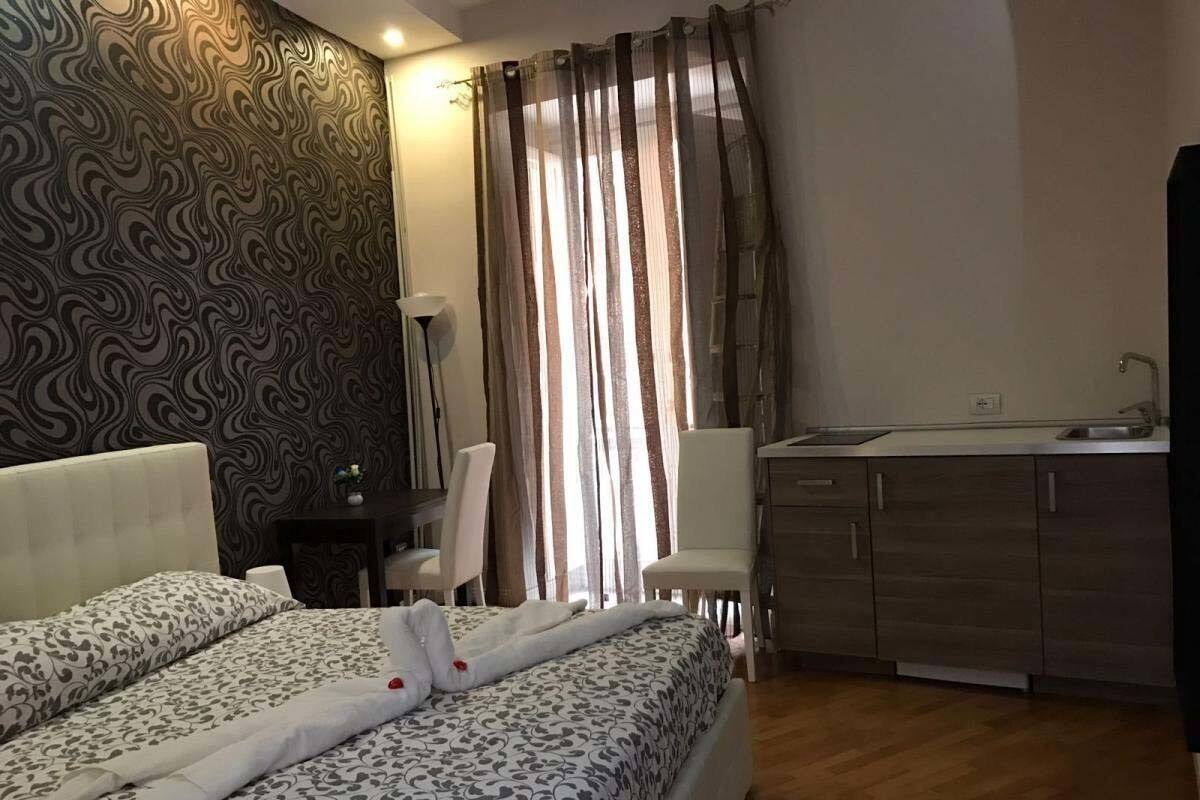 A hotel room with a double bed, a desk with a chair, a kitchenette, and a window with curtains.