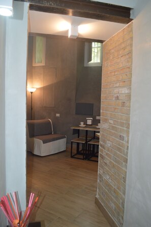 Un pasillo estrecho con pared de ladrillo a la derecha, suelo de madera, una pequeña mesa con sillas, una lámpara y un cuadro en la pared.