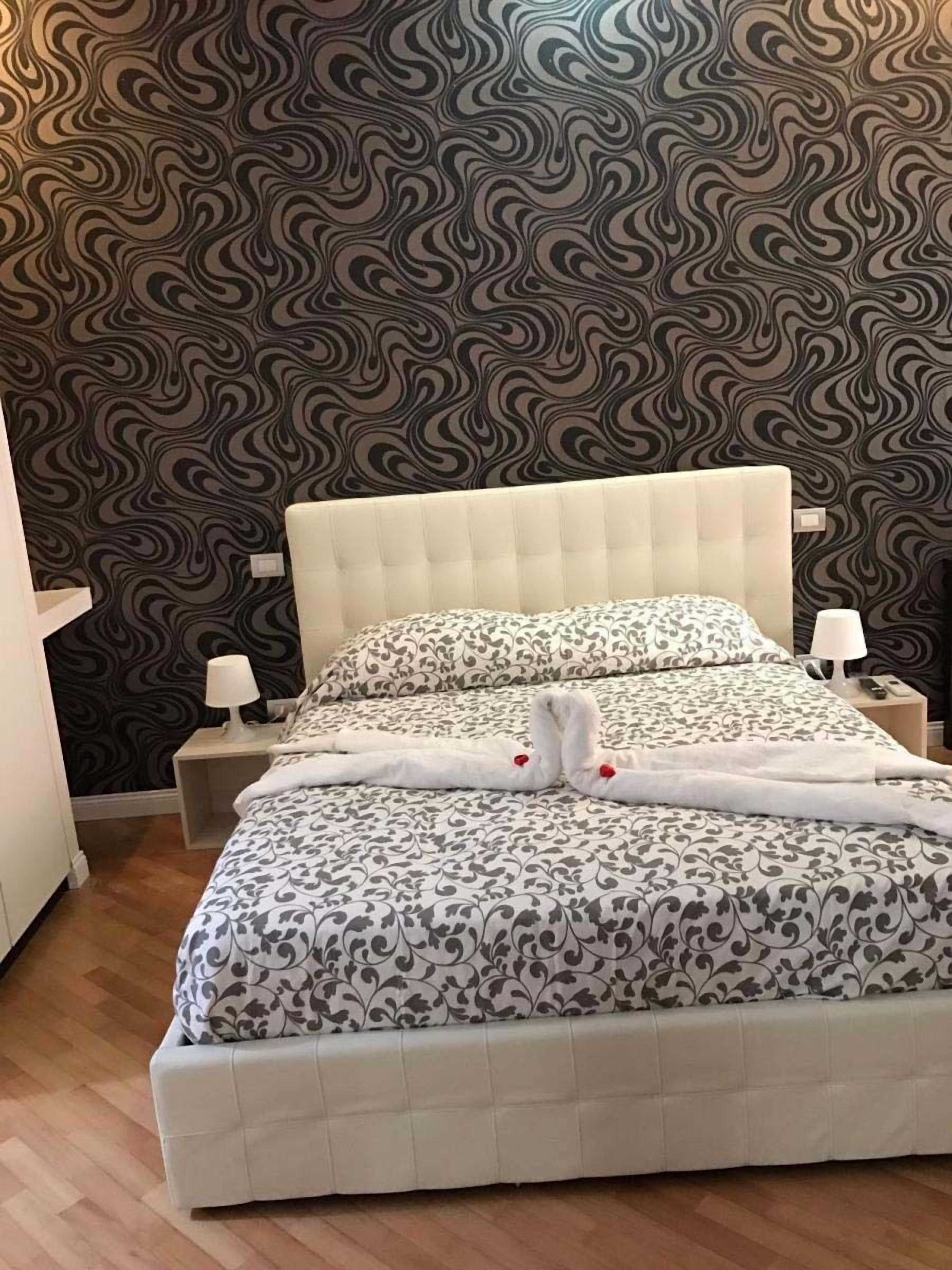 Un lit bien fait, recouvert d’une couvre-lit à motifs et agrémenté d’oreillers, flanqué de tables de chevet avec des lampes, dans une pièce dont le mur est recouvert d’un papier peint à motifs.