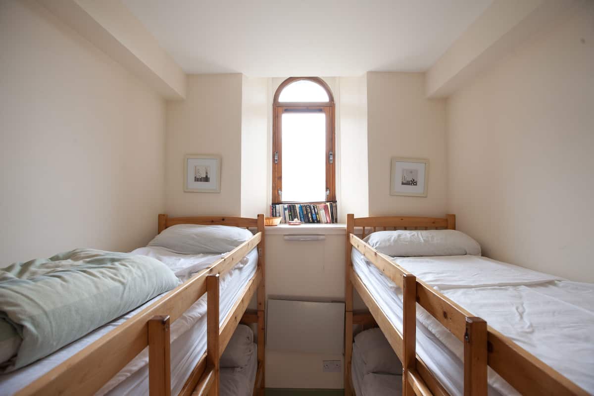 A small, narrow room with two wooden bunk beds, a window with a view, and a shelf with books.