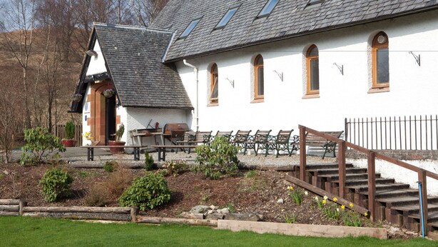A building with a sloped roof, multiple windows, and a staircase leading to a porch area with outdoor seating.