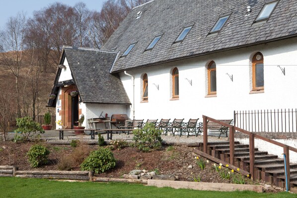 A building with a sloped roof, multiple windows, and a staircase leading to a porch area with outdoor seating.