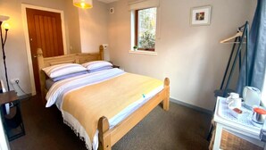 A single-bed room with a wooden frame, a bedside table with a lamp, a window with a view, and a door.