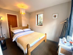 A single-bed room with a wooden frame, a bedside table with a lamp, a window with a view, and a door. - Inversnaid Bunkhouse - Hostel (Arrochar)