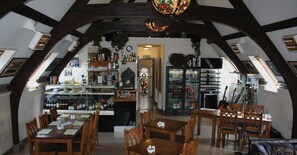 A cozy, rustic interior with wooden beams and a large, ornate chandelier. There are tables and chairs arranged for dining, and a variety of bottles and glasses on shelves.
