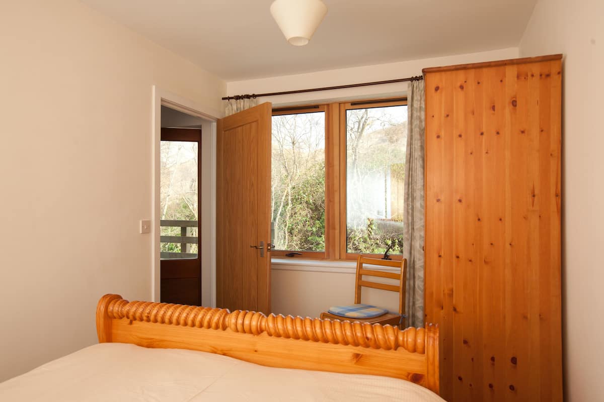 A bedroom with a wooden bed, a window with curtains, a wooden door, and a chair.