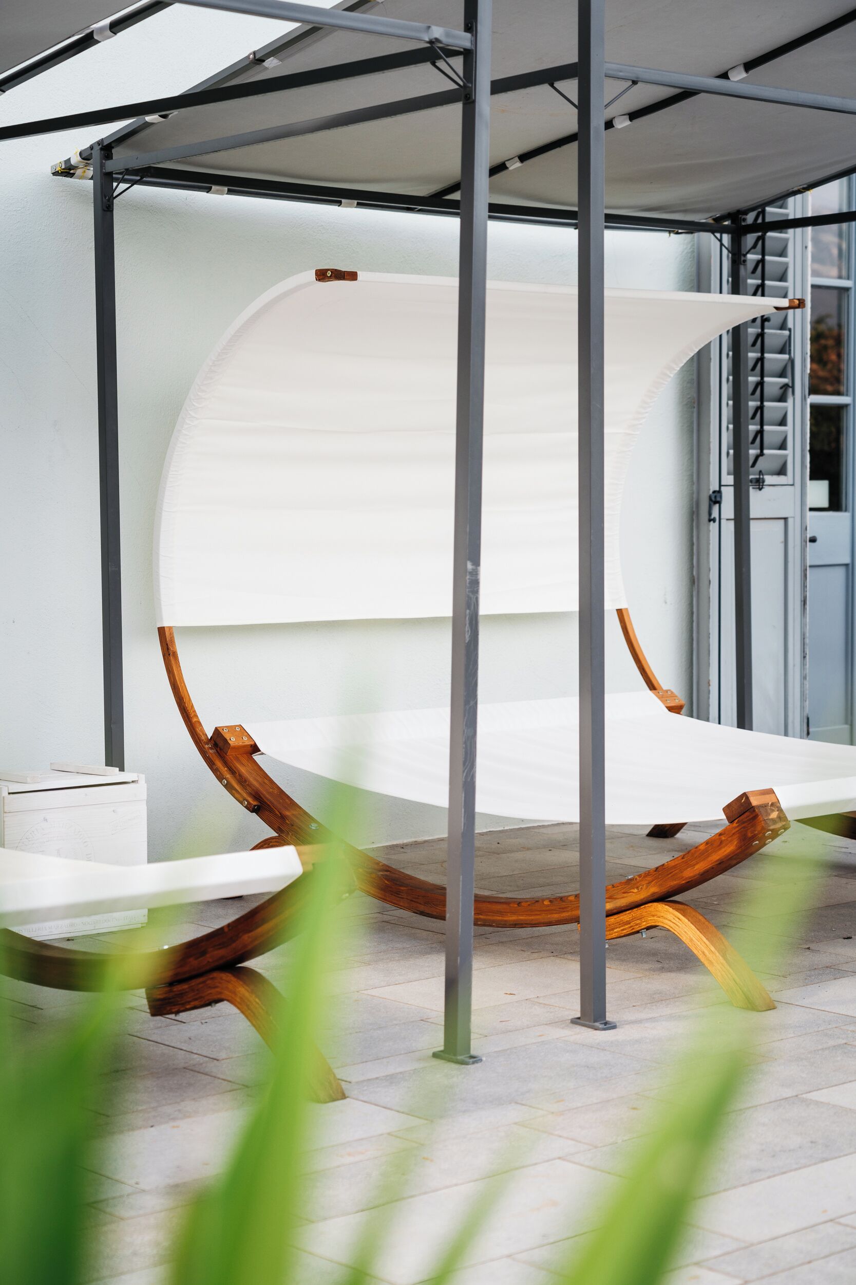 Un fauteuil suspendu, de type hamac, est placé sous une structure métallique surmontée d’une toile blanche, le tout adossé à la façade d’un bâtiment.