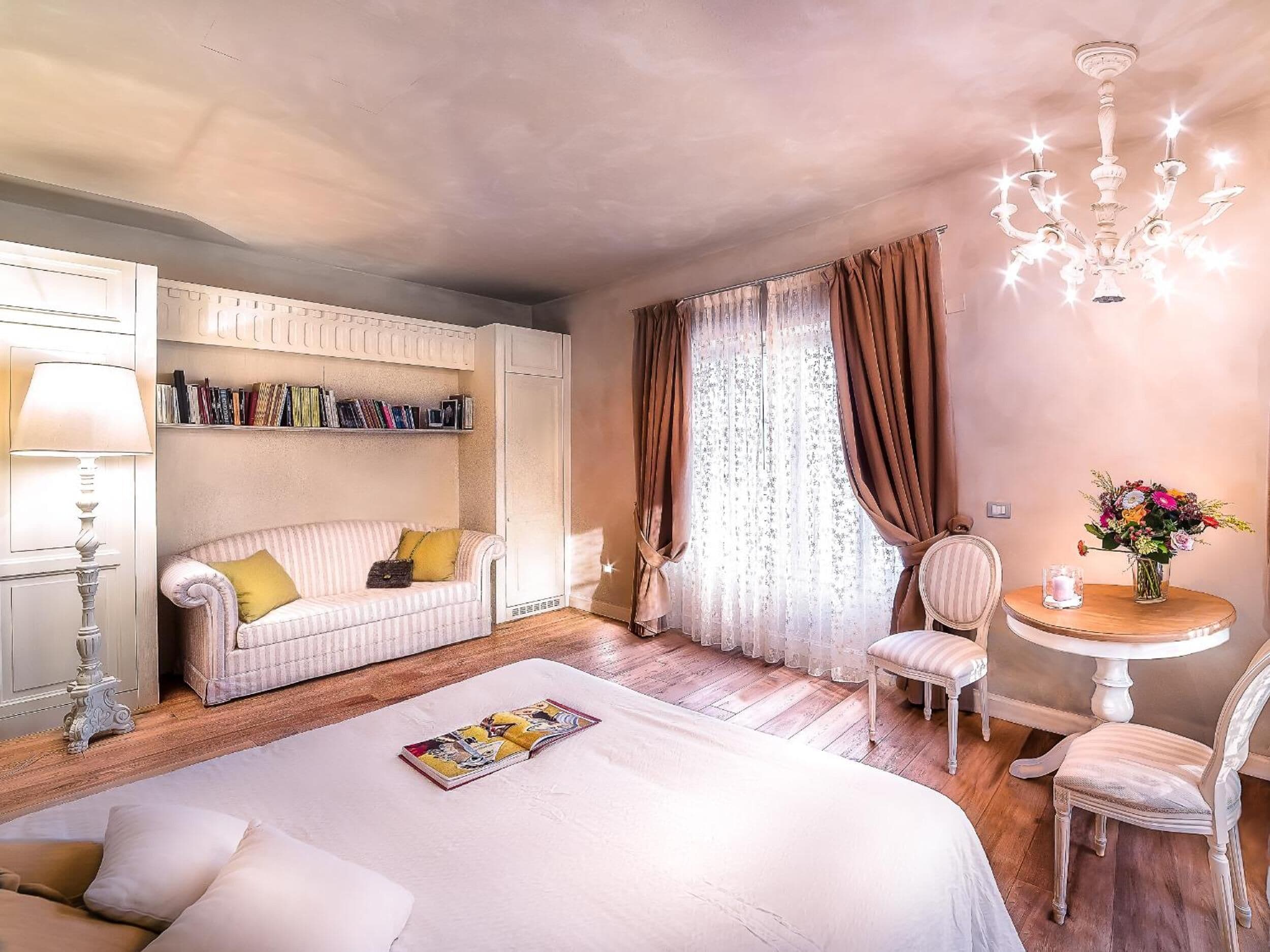 Une chambre d’hôtel confortable, avec un lit, un fauteuil de lecture, une petite table agrémentée d’un vase de fleurs, une bibliothèque et un lustre.
