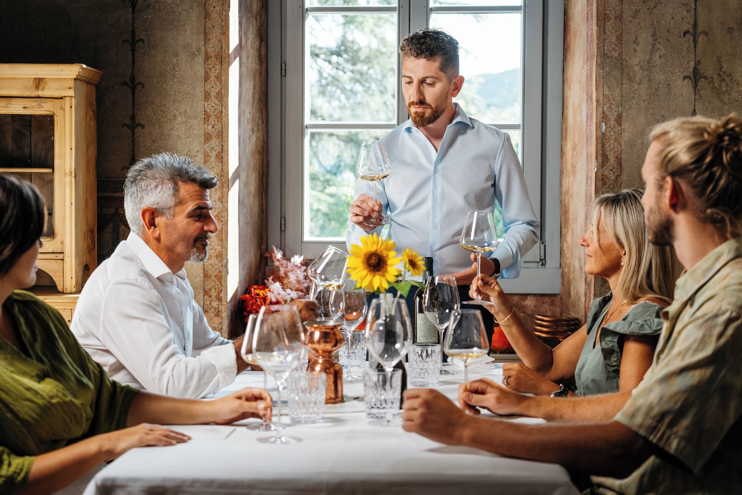 A group of people seated around a table with wine glasses, a vase with sunflowers, and a wooden cabinet in the background.