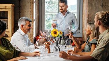 A group of people seated around a table with wine glasses, a vase with sunflowers, and a wooden cabinet in the background.