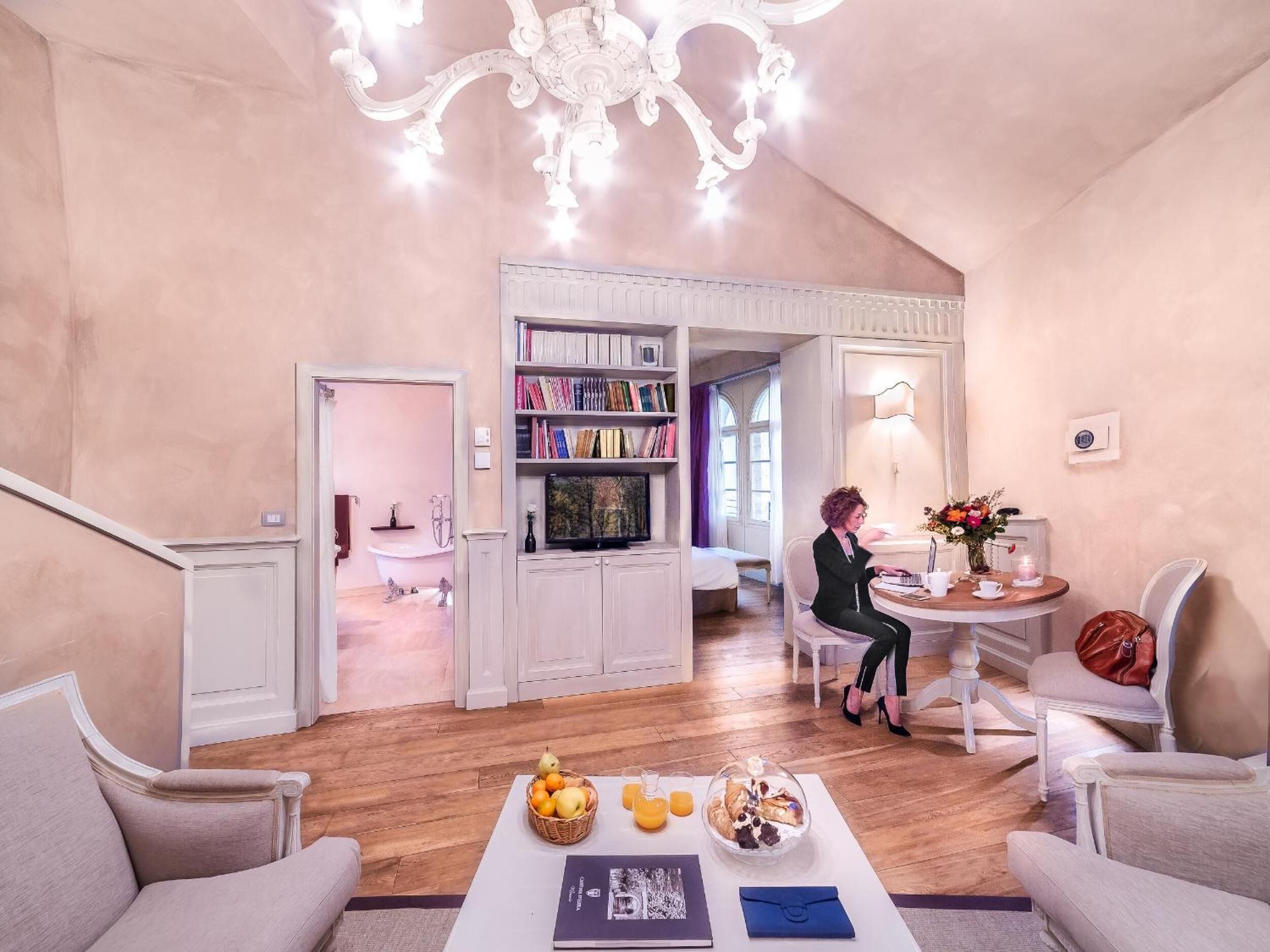 A spacious living room with a high ceiling, a chandelier, a bookshelf, a dining area with a table set for two, and a cozy sitting area with a sofa and armchairs.