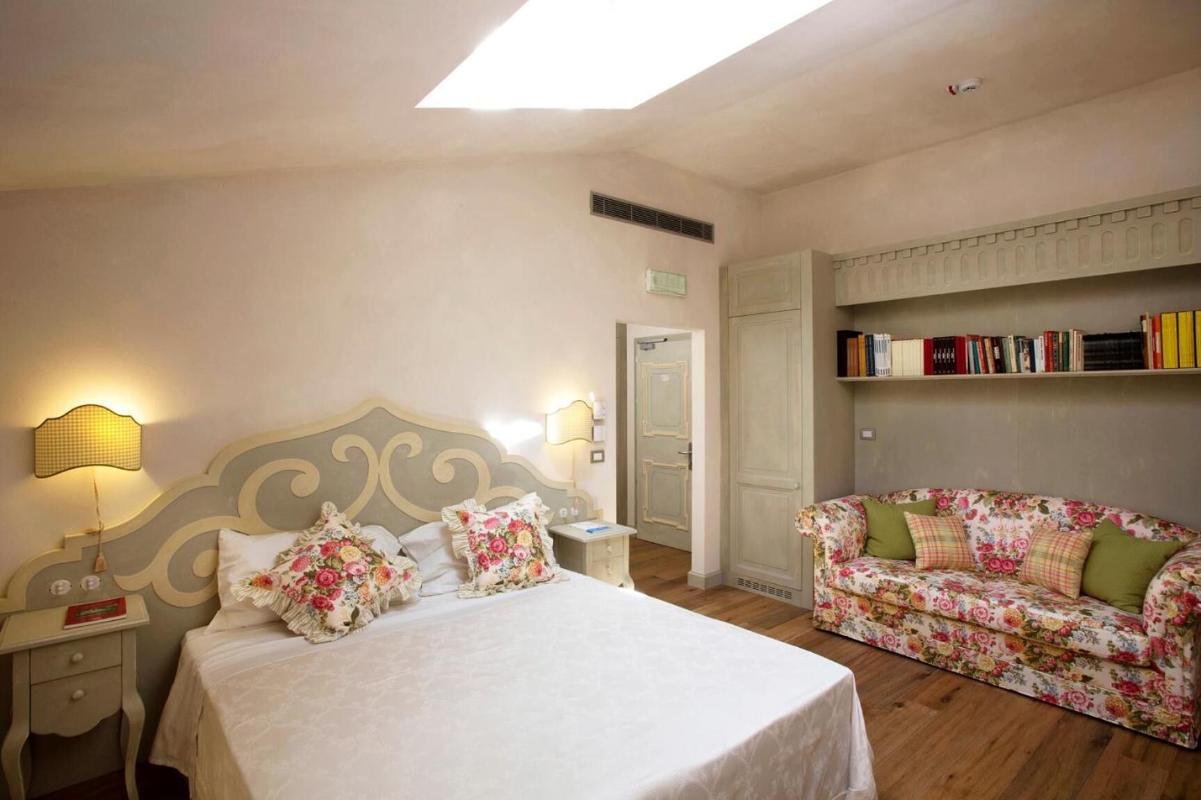 A bedroom with a bed, bedside lamps, a floral sofa, a bookshelf, and a door.